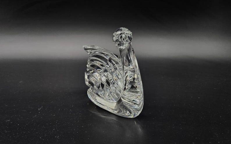 May include: A clear glass swan figurine with intricate detailing. The swan is positioned on a dark surface, showcasing its elegant form. The design features a detailed head and a textured wing, creating a decorative piece.