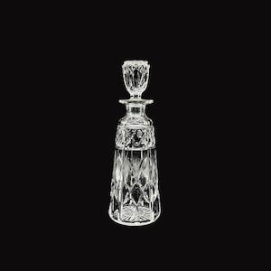 May include: A clear glass decanter featuring a faceted stopper and a body adorned with a geometric diamond-cut pattern. The base of the decanter displays a starburst design, while the neck is smooth and cylindrical, leading to a flared rim.