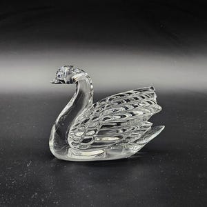 May include: A clear glass swan figurine with detailed wing and feather patterns. The swan is positioned on a dark surface, showcasing its elegant form. This decorative piece is ideal for home decor.