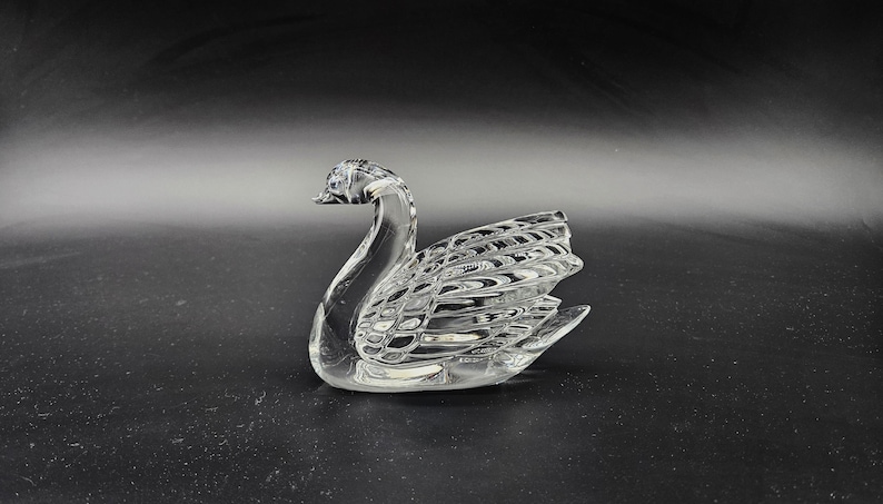 May include: A clear glass swan figurine with detailed wing and feather patterns. The swan is positioned on a dark surface, showcasing its elegant form. This decorative piece is ideal for home decor.