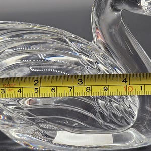 May include: A clear glass swan figurine, approximately 5 inches long, with detailed wing and feather patterns. The swan is measured with a yellow tape measure, showcasing its size and craftsmanship. The figurine is a decorative item.