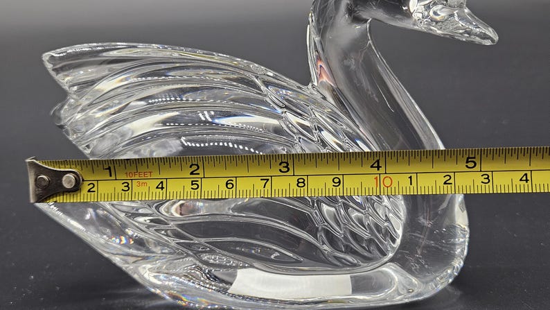 May include: A clear glass swan figurine, approximately 5 inches long, with detailed wing and feather patterns. The swan is measured with a yellow tape measure, showcasing its size and craftsmanship. The figurine is a decorative item.