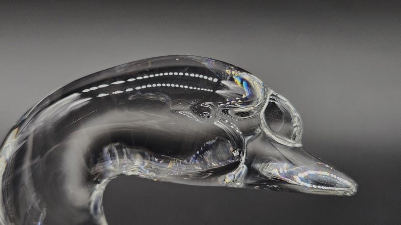May include: A clear glass sculpture of a swan's head and neck. The swan's head is detailed with a beak and eye, and the neck curves gracefully. The glass has a smooth, reflective surface, with a few air bubbles.