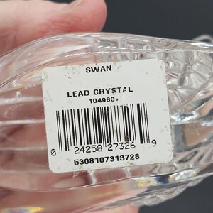 May include: A clear lead crystal swan figurine. The label reads "SWAN LEAD CRYSTAL" with a barcode and numbers. The swan has a textured, ribbed design. The figurine is held by a hand.