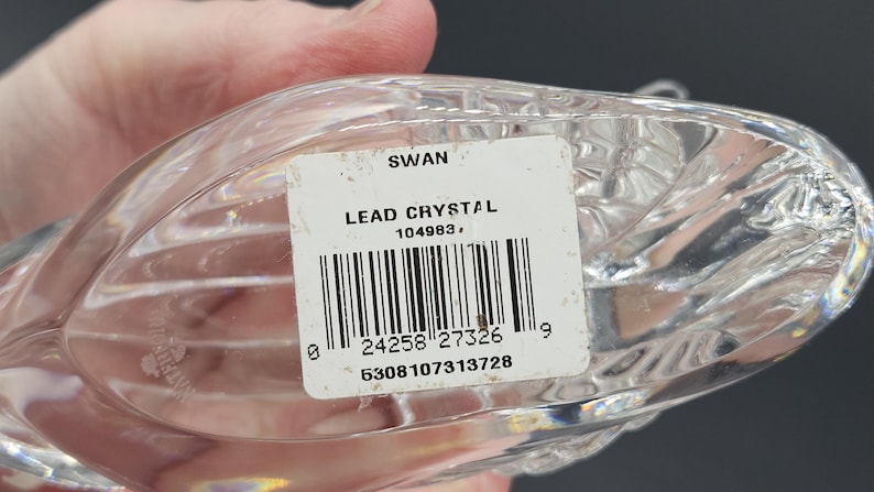 May include: A clear lead crystal swan figurine. The label reads "SWAN LEAD CRYSTAL" with a barcode and numbers. The swan has a textured, ribbed design. The figurine is held by a hand.