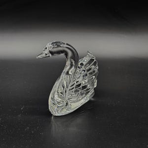 May include: A clear glass swan figurine with intricate detailing. The swan is positioned on a dark surface, its neck gracefully curved. The wings and body feature a textured pattern, reflecting light.