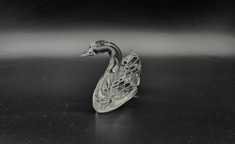 May include: A clear glass swan figurine with intricate detailing. The swan is positioned on a dark surface, its neck gracefully curved. The wings and body feature a textured pattern, reflecting light.