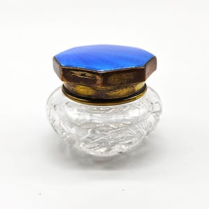 May include: A small, clear glass jar with a textured pattern. The jar has a brass-coloured rim and a blue, hexagonal lid. The lid has a metallic border. The jar is likely a vintage perfume bottle.
