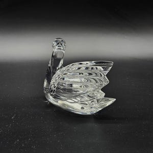 May include: A clear glass swan figurine with detailed wing and body textures. The swan is positioned on a dark surface, showcasing its elegant form. This decorative piece is ideal for home decor.
