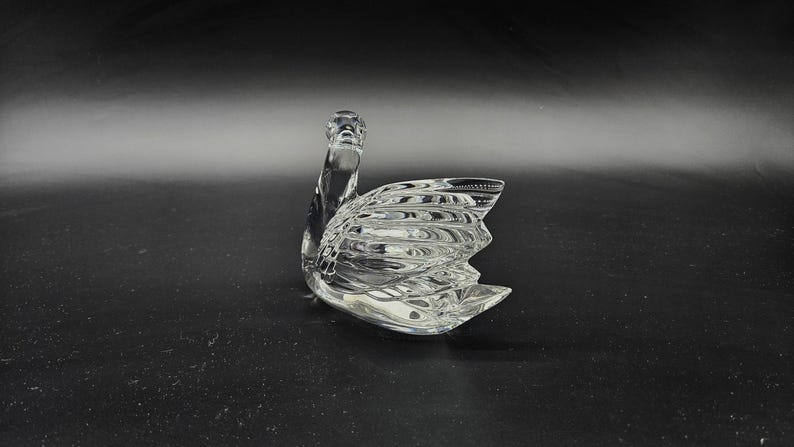 May include: A clear glass swan figurine with detailed wing and body textures. The swan is positioned on a dark surface, showcasing its elegant form. This decorative piece is ideal for home decor.