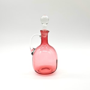 May include: A pink glass decanter with a clear glass stopper and a white handle. The decanter has a rounded body and a narrow neck. The stopper is a small, clear sphere. The handle is a curved, white glass. The decanter is empty.