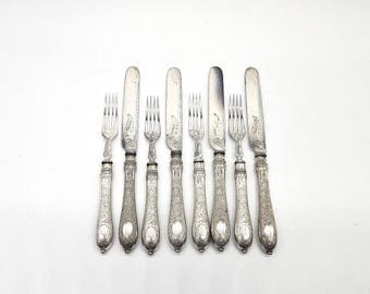 May include: A set of antique silverware, including forks and knives, arranged on a white surface. The silver utensils feature ornate, engraved handles and blades. The arrangement includes four forks and four knives.