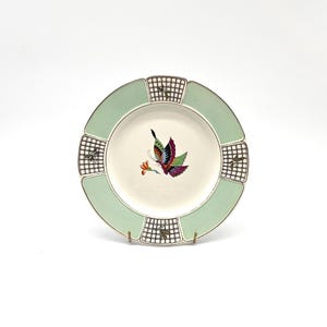 May include: A decorative plate with a pale green border and a cream-coloured centre. The plate features a colourful butterfly design in the centre. The border has a gold trim and chequered accents. The plate is displayed on a gold stand.