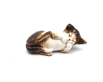 May include: A ceramic figurine of a cat lying on its back. The cat has brown and white markings, with its paws and tail curled. The cat's eyes are closed, and it appears to be sleeping or relaxed.
