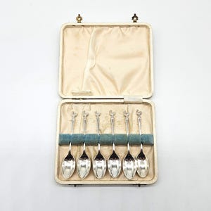 May include: A set of six silver spoons presented in a cream-coloured case. The spoons feature decorative handles and are arranged in a blue velvet-lined compartment. The case is open, displaying the spoons against a cream satin interior.