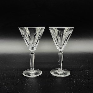 May include: A pair of clear crystal wine glasses with an etched pattern. Each glass has a stemmed base and a V-shaped bowl. The background is black, accentuating the shape and clarity of the glassware.