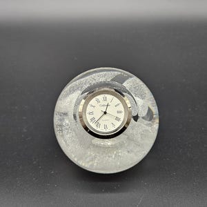 Può includere: Un orologio fermacarte sferico in vetro trasparente con quadrante bianco, numeri romani neri e la scritta "Cottage" stampata in nero. Il corpo in vetro presenta un aspetto testurizzato e satinato con motivi vorticosi interni e una lunetta argentata.