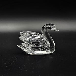 May include: A clear glass swan ornament with intricate feather and wing detailing. The swan is set against a dark background, highlighting its graceful shape. A decorative item suitable for home display.