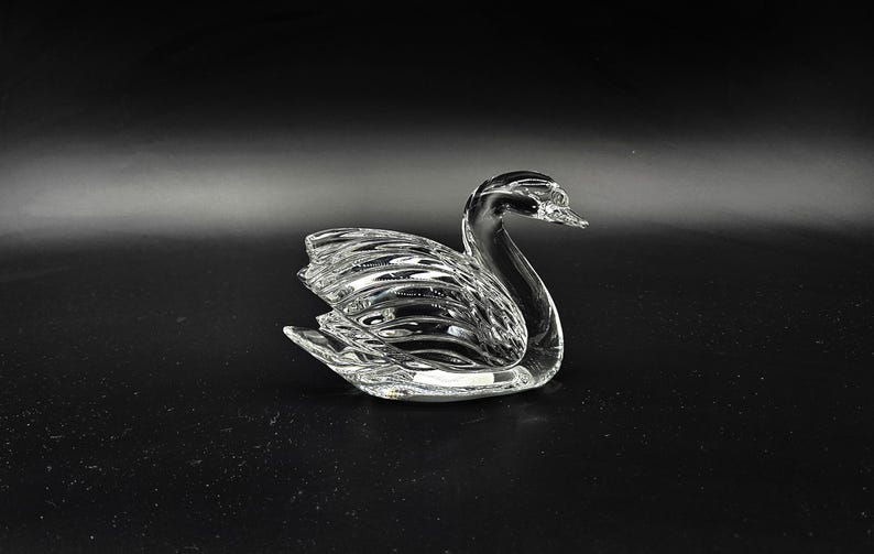 May include: A clear glass swan figurine with detailed feather and wing textures. The swan is positioned on a dark surface, showcasing its elegant form. This decorative piece is ideal for home decor.