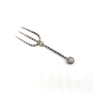 Op de afbeelding: Zilveren cocktailvork in de vorm van een drietand, met een gedraaide steel en decoratieve kogelvormige uiteinden. De drie tanden zijn scherp en de steel heeft een textuur. De vork staat tegen een witte achtergrond.