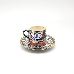 Peut inclure: Une petite tasse à expresso en céramique avec sa soucoupe assortie, présentant une finition irisée sombre ornée d'un motif floral multicolore vif. Le bord de la tasse et celui de la soucoupe sont soulignés par un fin liseré doré métallique.