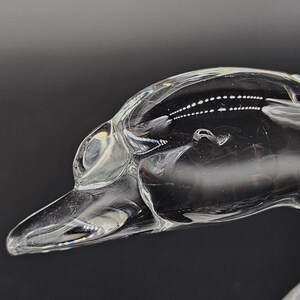 May include: A clear glass sculpture of a duck's head and neck, with a long beak and detailed eye. The sculpture is transparent, reflecting light and set against a dark background. The artwork is a decorative item.