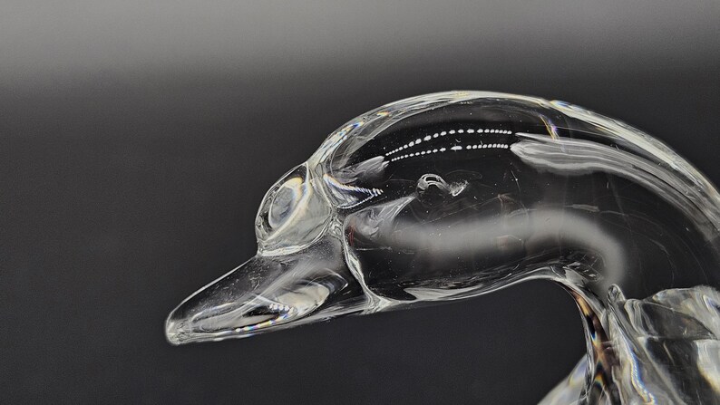 May include: A clear glass sculpture of a duck's head and neck, with a long beak and detailed eye. The sculpture is transparent, reflecting light and set against a dark background. The artwork is a decorative item.