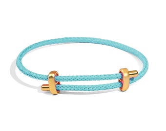 May include: A turquoise braided bracelet with gold-colored metal accents. The bracelet features a double-strand design with cylindrical gold clasps. The bracelet is a stylish accessory.