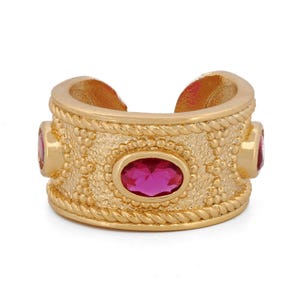 May include: Gold-toned cuff ring with three oval, magenta-colored gemstones. The ring features a textured band with a rope-like border and a central oval gemstone. The design is ornate and detailed, suitable for jewelry.