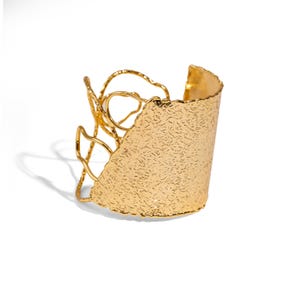 May include: A gold-colored cuff bracelet with a textured surface and an open, abstract design on one side. The bracelet has a wide band and a decorative, organic shape.