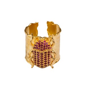 May include: Gold ring with a textured band and a pink beetle design. The beetle is covered in small pink rhinestones.
