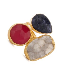 May include: Three gold-toned rings with large, colorful gemstones. One ring features a faceted red stone, another a dark blue teardrop-shaped stone, and the third a white, textured stone. The rings are set against a white background.