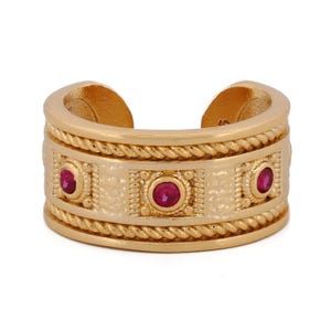 May include: Gold-tone ring with three red gemstones. The ring features a textured band with a rope-like design on the top and bottom edges. The gemstones are set in square frames.