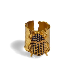 May include: A gold ring with a textured band and a blue beetle design. The beetle is made of small blue stones.