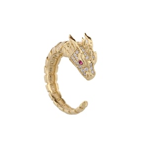 Puede incluir: Un anillo de tono dorado con un diseño de dragón. La cabeza del dragón está adornada con piedras de circonita cúbica transparentes y rojas.