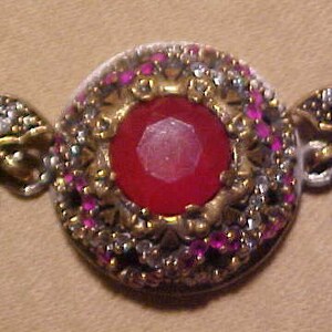 May include: A silver and gold bracelet with a large red gemstone surrounded by smaller pink and white gemstones.