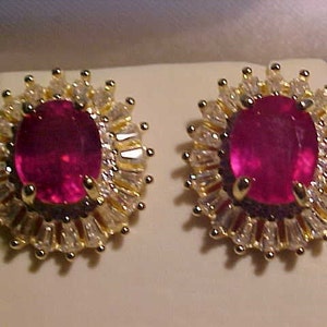 May include: A pair of gold earrings featuring a large oval ruby gemstone surrounded by smaller clear gemstones.