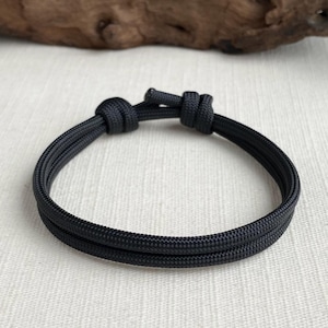 May include: Black paracord bracelet with a sliding knot closure. The bracelet is made of two parallel strands of black cord, creating a simple, minimalist design. The bracelet is shown on a neutral background.