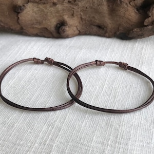 May include: Two brown cord bracelets tied in a knot. The bracelets are on a white fabric background.