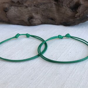 May include: Two green string bracelets with adjustable knots. The bracelets are made of thin, round, green cord. The knots are centered on the bracelets. The background is a light-colored fabric and a piece of driftwood.