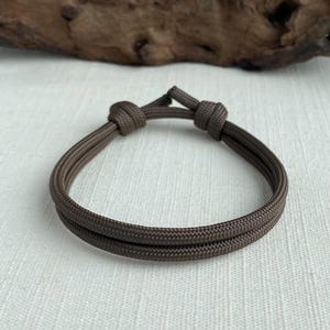 May include: A brown adjustable bracelet made of woven cord. The bracelet features two parallel strands and is secured with two knots. The background is a light, textured surface with a piece of wood in the upper portion.