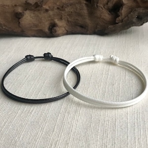 May include: Two friendship bracelets, one black and one white, made of thin cord, tied in a simple knot.