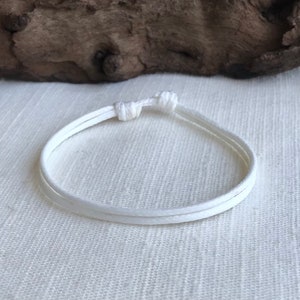 May include: A white bracelet made of three cords, knotted together. The bracelet is simple and minimalist, with a clean, bright appearance. The background is a light-colored, textured surface.