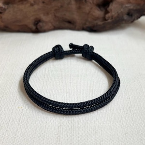May include: A black cord bracelet featuring a braided texture and a double-strand design. The bracelet is secured with two prominent knots at the top, creating a simple and functional closure mechanism for this wrist accessory.