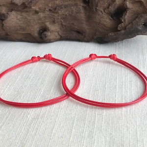 May include: Two red string bracelets tied in a knot. The bracelets are on a white fabric background.