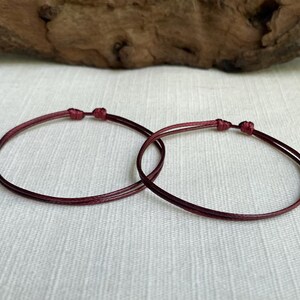 May include: Two burgundy-colored string bracelets with adjustable knots. The bracelets are circular and lie on a white, textured surface. A piece of driftwood is in the background.
