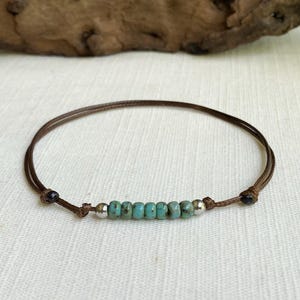 May include: A brown cord bracelet featuring a row of turquoise beads and silver accents. The bracelet is knotted at each end with a small black bead. The jewelry is displayed on a white surface.