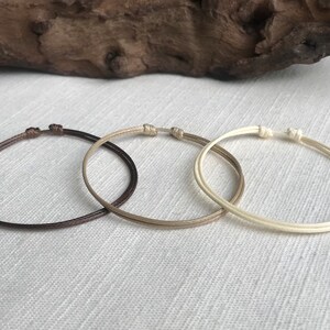 May include: Three simple bracelets made of thin, braided cord. The bracelets are brown, beige, and white.