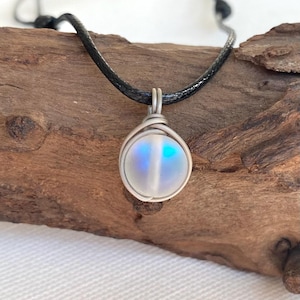 May include: A handcrafted pendant necklace featuring a round, iridescent stone encased in a silver wire wrap. The stone displays a spectrum of colors, including blue and white. The necklace has a black cord.