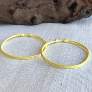 May include: Two delicate, adjustable bracelets in a soft yellow hue. Each bracelet features a double-strand design and a sliding knot closure. The bracelets are displayed on a white fabric background, with a piece of driftwood in the background.